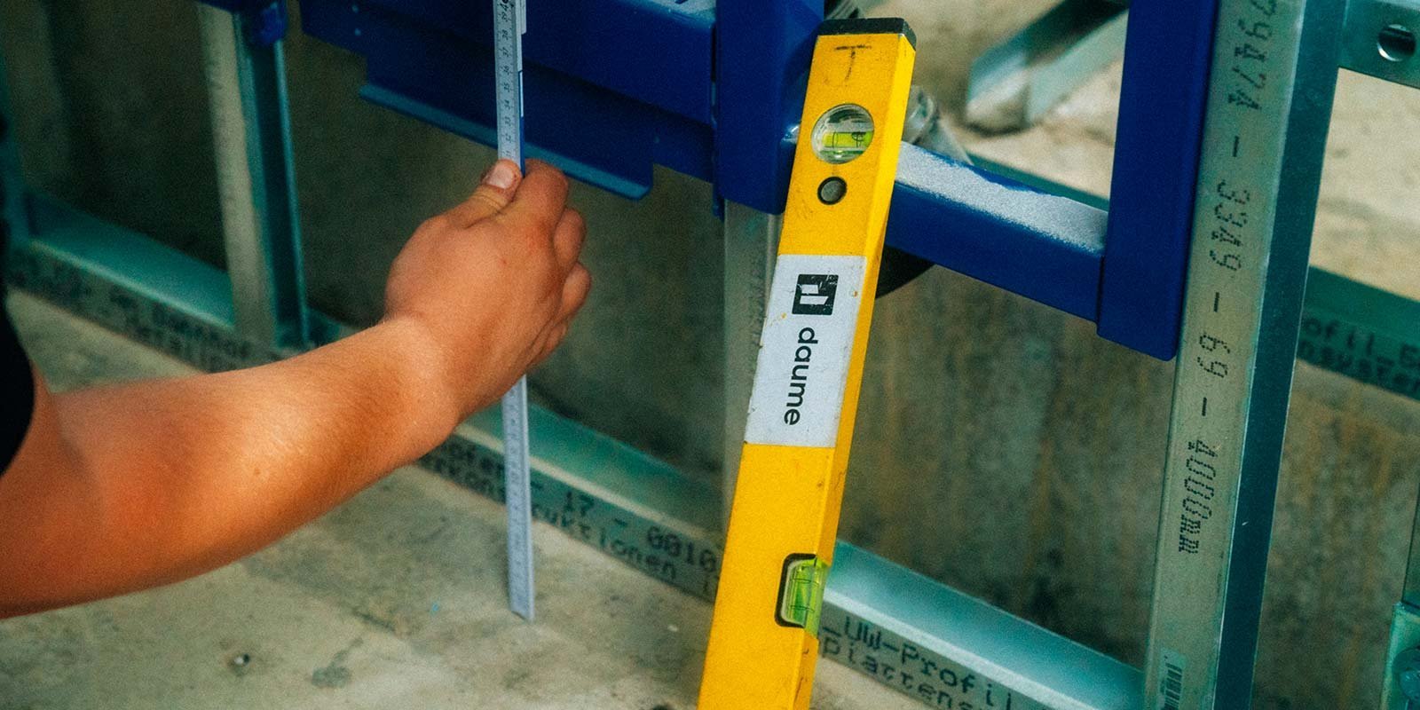 Der Arbeiter misst mit einem Maßstab an einer blauen Konstruktion, während eine gelbe Wasserwaage zur Kontrolle der Ebenheit verwendet wird.