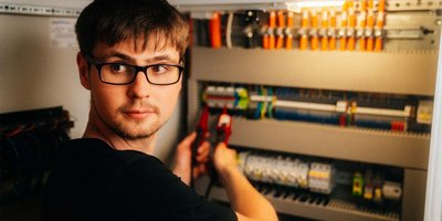 Ein junger Mann mit Brille arbeitet konzentriert an einem elektrischen Schaltschrank und hält eine Zange in der Hand.