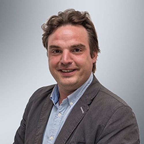 Ein lächelnder Mann mit lockigem Haar trägt einen grauen Blazer und ein hellblaues Hemd vor einem neutralen Hintergrund.