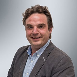 Ein lächelnder Mann mit lockigem Haar trägt einen grauen Blazer und ein hellblaues Hemd vor einem neutralen Hintergrund.