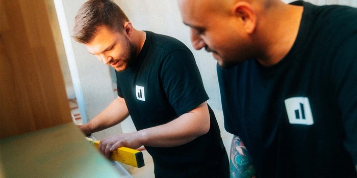 Zwei Männer in schwarzen T-Shirts arbeiten konzentriert an einem Projekt, einer hält ein Maßband, während der andere zuschaut.