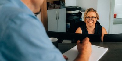 Im Büro lächelt eine blonde Frau mit Brille, während ein Mann am Tisch Notizen auf einem Block macht.