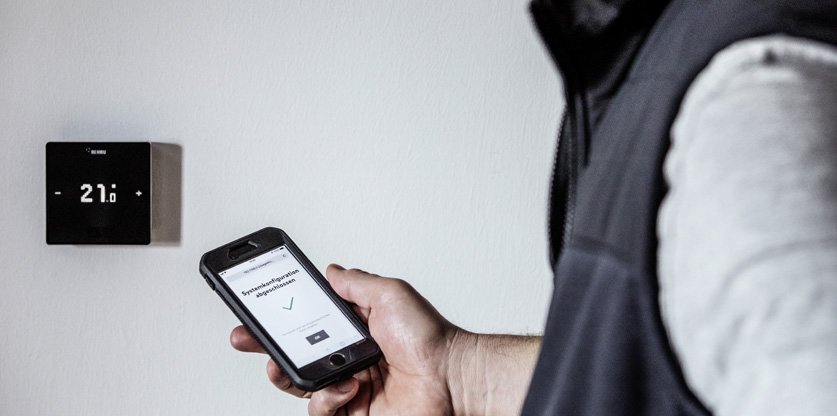 Eine Heizungssteuerung an der Wand und ein Mann der ein Handy in der Hand haelt, geoeffnet mit einer Steuerungs App 