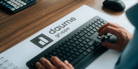 Eine Person nutzt eine schwarze Tastatur, während das Logo der Daume Gruppe auf einem weißen Blatt sichtbar ist.