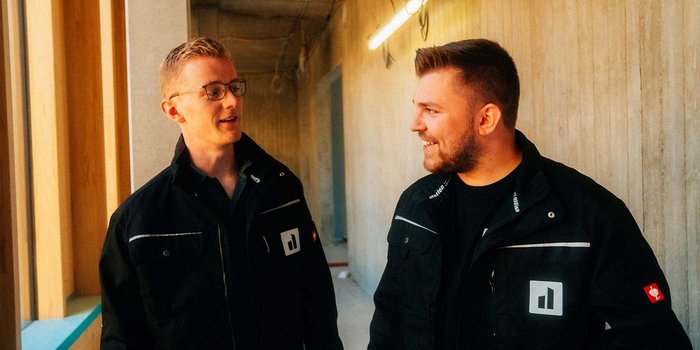 In einem Bauumfeld stehen zwei Männer in schwarzen Jacken und tauschen freundliche Blicke und Worte aus.