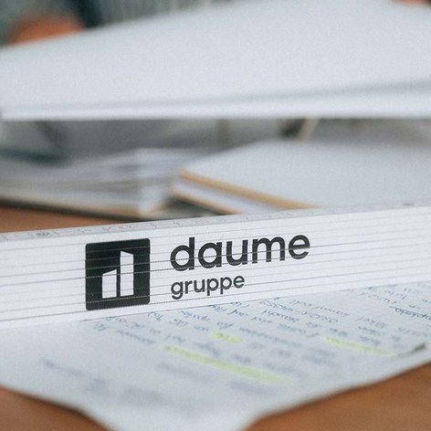 Der Fokus liegt auf einem weißen Block mit dem Logo der Daume Gruppe, umgeben von Notizen und einem Kaffeebecher.