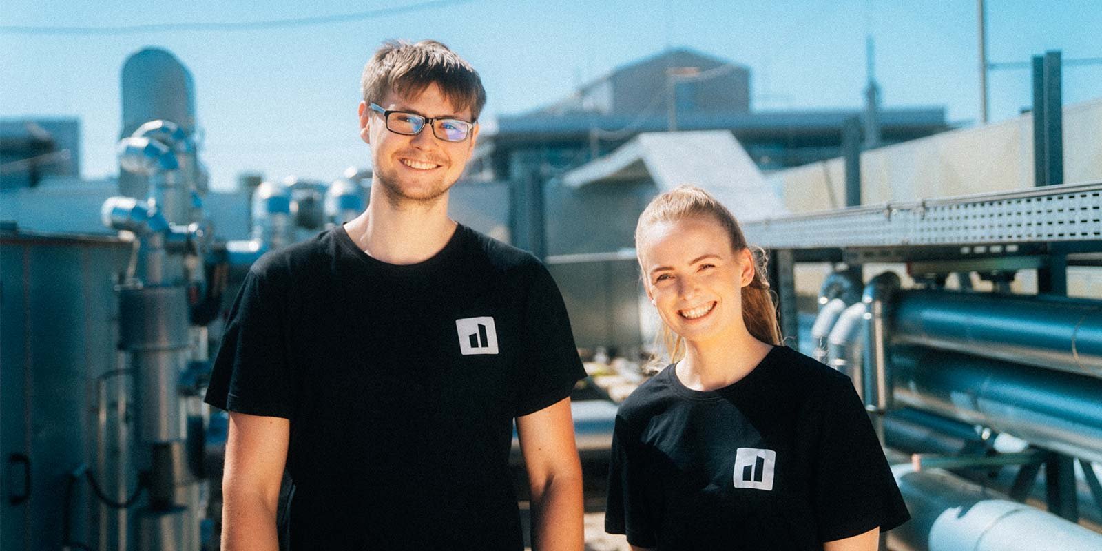 Zwei junge Menschen posieren fröhlich im Freien, umgeben von metallischen Anlagen und Rohren, die im Hintergrund sichtbar sind.