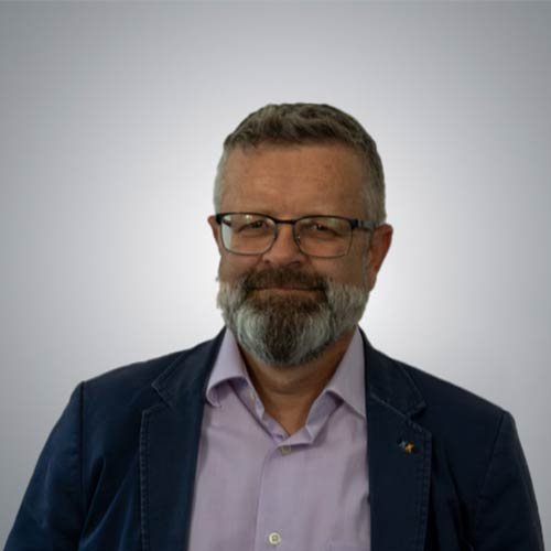 Ein Mann mit Brille und grauem Bart lächelt freundlich vor einem hellen Hintergrund.