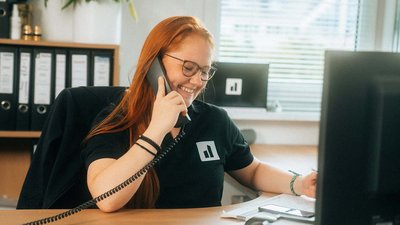 Die Mitarbeiterin mit Brille und rotem Haar telefoniert fröhlich, umgeben von Aktenordnern und einem Computerbildschirm.